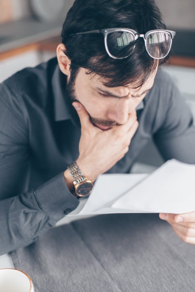 Man in gray shirt with glasses looking concerned over financial papers.