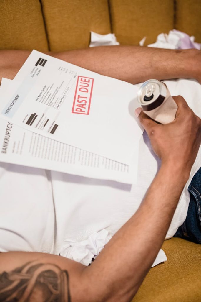 A man holding a drink lies on a couch with past due bills, feeling stressed.